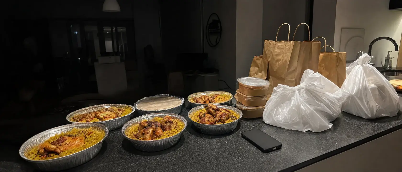 A dark kitchen counter lined with foil takeout trays of chicken and rice, paper bags and plastic delivery bags