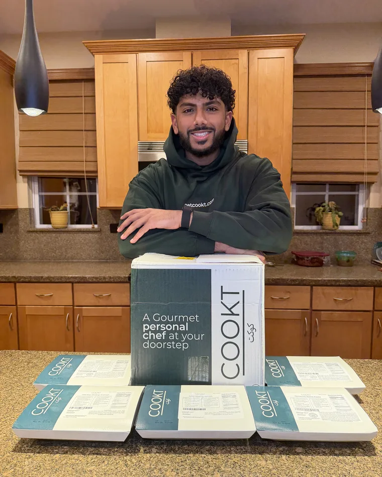 Farzy standing behind a COOKT delivery box and a row of sealed COOKT meals