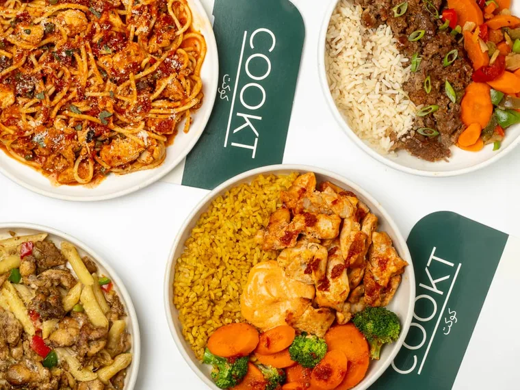 Four COOKT halal meals plated beside COOKT-branded meal sleeves