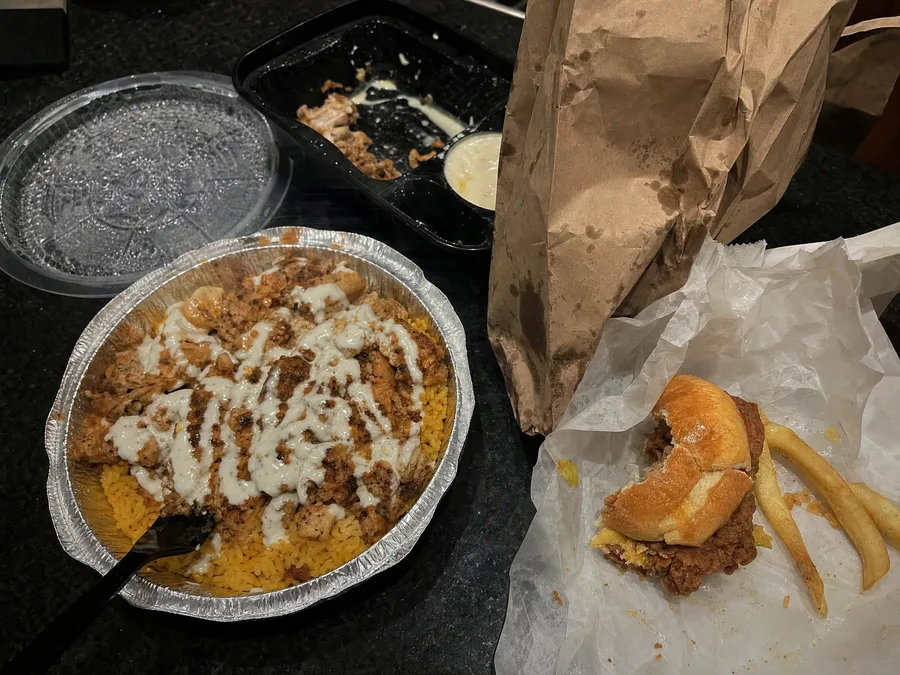 Greasy late night halal takeout on a dark kitchen counter: chicken over yellow rice in a foil tray, a fried chicken sandwich and fries