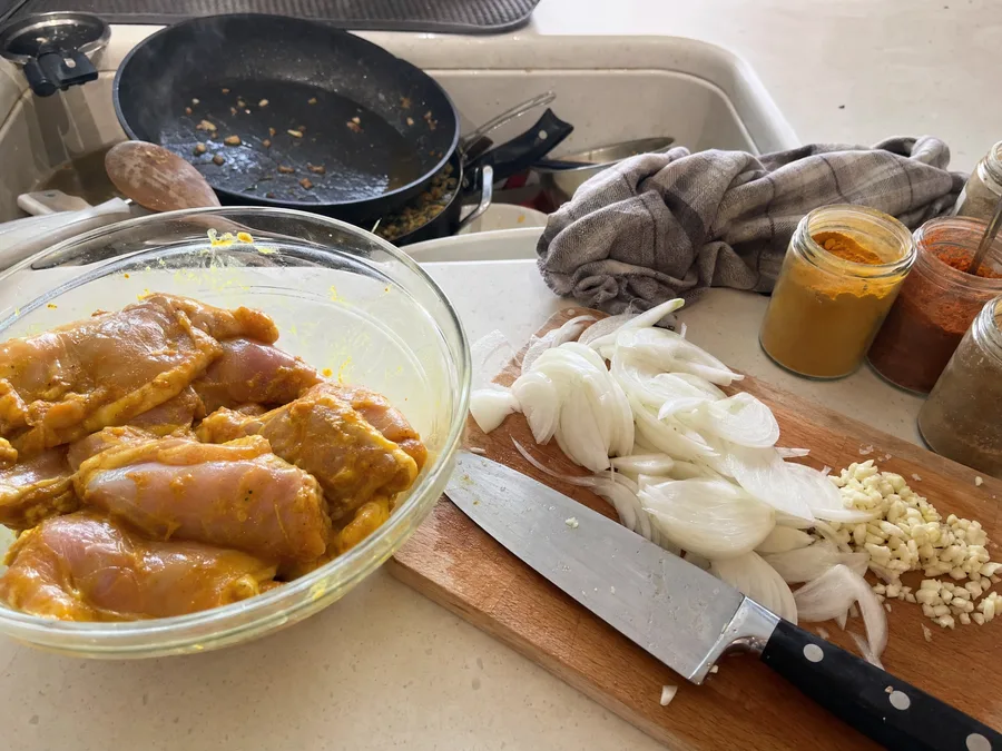 A home kitchen counter mid-cook with marinated chicken in a bowl, sliced onions and garlic on a board and a used pan in the sink